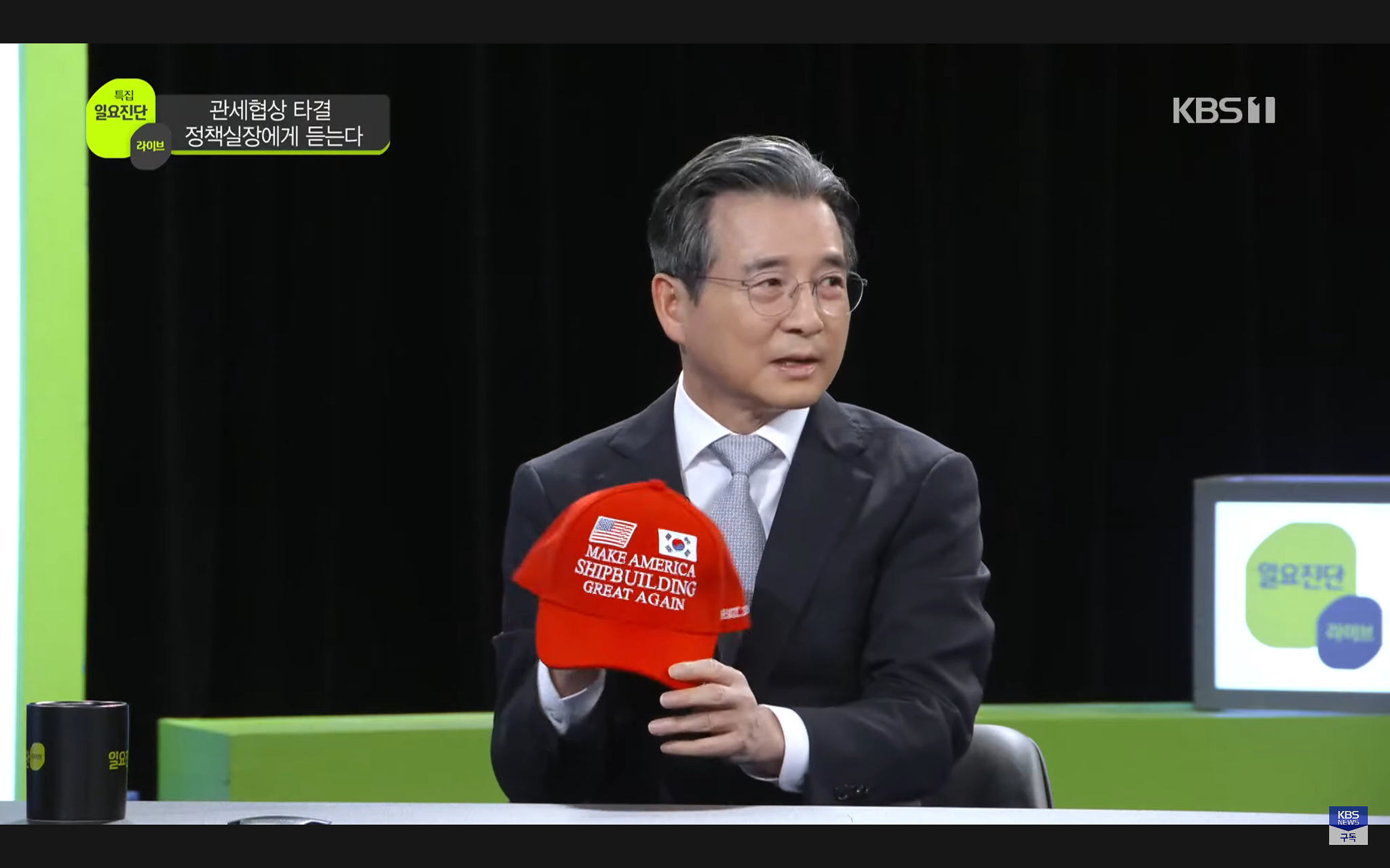 Kim Yong-beom, the presidential chief of staff for policy, presents a red baseball cap reading “Make America Shipbuilding Great Again” on KBS on Aug. 3. [SCREEN CAPTURE]